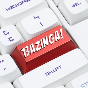 Peut inclure: Une touche de clavier rouge avec le mot "BAZINGA!" imprimé en lettres blanches. La touche est sur un clavier blanc.