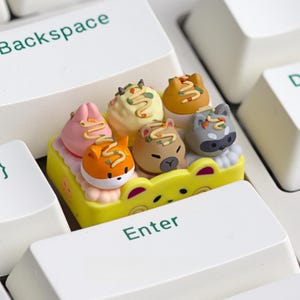 May include: A collection of colorful animal-themed keycaps, including a fox, bear, and raccoon, sits on a yellow tray. The keycaps are placed on a white keyboard, with the "Enter" and "Backspace" keys visible.