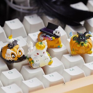 May include: A collection of Halloween-themed keyboard keycaps. The keycaps feature various designs, including a pumpkin with a bat, a ghost, a pumpkin with a hat and candy, and a pumpkin with a candle and a tombstone. The keycaps are orange, white, black, and purple.