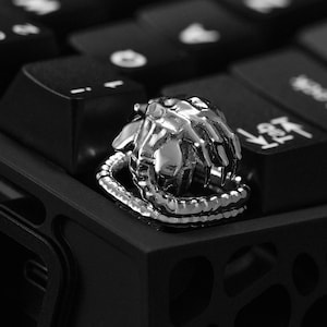 Puede incluir: Un anillo plateado con un diseño de mano esquelética, con detalles de huesos de los dedos y una banda texturizada. El anillo se encuentra sobre un teclado negro, creando un contraste llamativo.