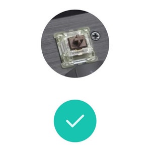 May include: A close-up of a mechanical keyboard switch. The switch has a clear plastic housing with a brown stem and a metal contact. The switch is mounted on a gray surface with a screw visible.