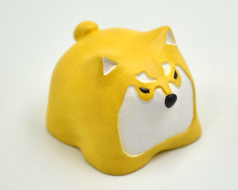 Op de afbeelding: Een geel keramiek beeldje van een Shiba Inu hond met witte gezichtsmarkeringen en zwarte ogen. De hond zit met zijn hoofd naar beneden gekanteld en zijn mond gesloten.