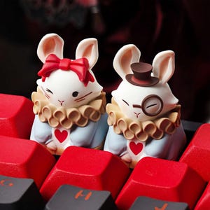 Può includere: Due statuette di conigli bianchi, una con un fiocco rosso, l'altra con un piccolo cappello marrone e un monocolo. Entrambi hanno colletti arruffati, giacche blu e dettagli a cuore rosso. Sono posizionati su una tastiera con tasti rossi.