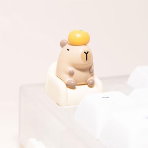 Peut inclure: Figurine de capybara beige avec un fruit jaune sur la tête, assis sur une petite chaise blanc cassé. La figurine porte un 'Y' noir sur la poitrine. Le fond est de couleur neutre.
