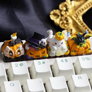 Può includere: Primo piano di una tastiera di computer con tasti personalizzati a tema Halloween. I tasti presentano zucche arancioni, fantasmi e un pipistrello. I tasti sono bianchi con lettere e simboli neri.