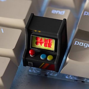 Könnte beinhalten: Ein schwarzer Miniatur-Arcade-Maschinen-Tastenkappe mit rotem Joystick, blauen und gelben Knöpfen und einem Bildschirm mit der Aufschrift "GAME OVER". Die Tastenkappe befindet sich auf einer weißen Tastatur.