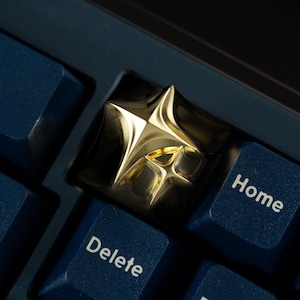 May include: A metallic gold artisan keycap featuring a four-pointed star design with sharp, raised edges. The keycap is installed on a dark blue mechanical keyboard between keys labeled Delete and Home in white sans-serif font.