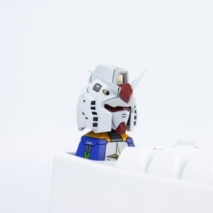 May include: A white and red robot head with a blue and yellow torso. The robot is partially obscured by a white surface.