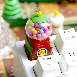 May include: A miniature gumball machine keycap with a clear globe filled with colorful candies. The base is red and green with a red bow. A yellow star sits atop a green leafy top. The keycap is placed on a white keyboard.