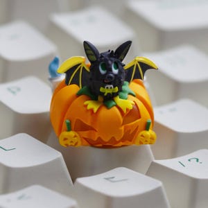 May include: A Halloween-themed keyboard keycap featuring a carved orange pumpkin with a smiling face. A black bat with yellow-trimmed wings sits atop the pumpkin. The keycap is surrounded by white keyboard keys.