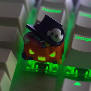 May include: A Halloween-themed artisan keycap featuring a jack-o'-lantern design with a black top hat and a ghost. The orange pumpkin has glowing green eyes and mouth. The keycap is on a keyboard with green backlighting.