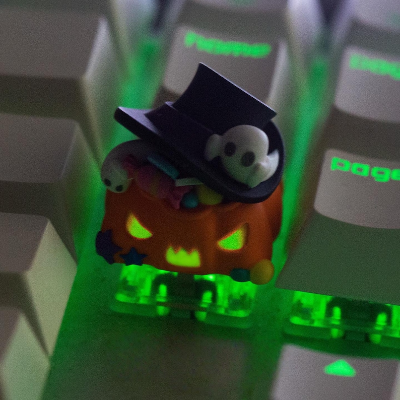 May include: A Halloween-themed artisan keycap featuring a jack-o'-lantern design with a black top hat and a ghost. The orange pumpkin has glowing green eyes and mouth. The keycap is on a keyboard with green backlighting.
