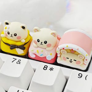 Puede incluir: Tres teclas decorativas con forma de oso en amarillo, rosa y blanco, apoyadas en un teclado blanco. Cada tecla presenta un diseño de oso diferente con expresiones faciales lindas y acentos coloridos. Las teclas están en las teclas 7, 8 y 9.