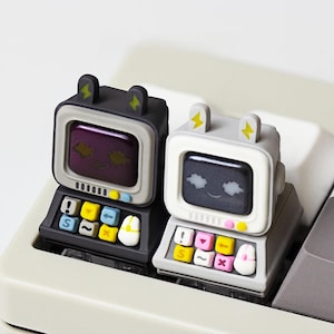 May include: Two mechanical keyboard keycaps shaped like miniature retro computers with cat ears. One is dark grey with a purple screen, the other is light grey with a black screen. Both feature small colourful keys and a mouse icon on the front.