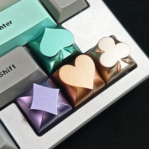 May include: Close-up of a mechanical keyboard with custom keycaps. The keycaps feature playing card suit symbols: spades, hearts, diamonds, and clubs. The keycaps have metallic finishes in various colors, including teal, gold, and purple. The keyboard has a silver frame.