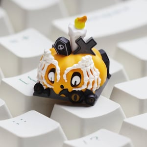 May include: A Halloween-themed artisan keycap featuring a pumpkin design with a skeleton face, a candle, and a tombstone. The keycap is orange, white, black, and yellow, and sits on a white keyboard.