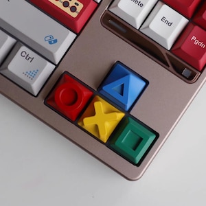 以下が含まれることがあります： カラフルなキーキャップを備えたカスタムメカニカルキーボード。 白、赤、グレーのキーが混在し、赤、青、黄、緑の鮮やかな幾何学模様のキーが配置されています。 キーボードはブロンズ色のフレームです。
