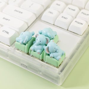 May include: A close-up of a white mechanical keyboard with custom keycaps. The keycaps are light blue elephant figurines sitting on green miniature couches. The keyboard keys are white.