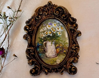 May include: An oval-shaped painting in a dark, ornate, metallic-toned frame featuring a textured depiction of a white pitcher filled with white daisies and green foliage. The artwork is displayed alongside dried floral stems and two small purple rosebuds.