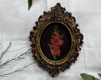 May include: An oval-shaped wall frame with an ornate, antique gold-colored finish featuring intricate scrollwork details. Inside the frame, a piece of black fabric displays a hand-stitched embroidery of a vibrant pink flower with green stems.