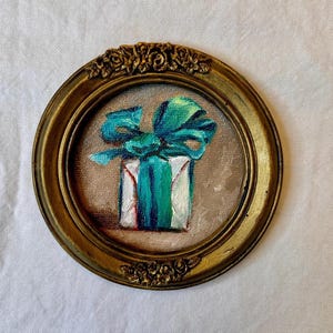 May include: A miniature painting of a gift box in a small, round, antique gold-coloured frame. The gift box is white with teal stripes and a teal bow. The painting is set against a beige background.