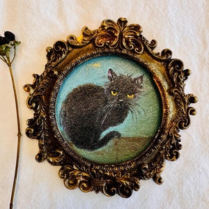 May include: A vintage-style framed print featuring a black cat with yellow eyes. The oval print is set in an ornate, gold-colored frame. A dried plant stem with black berries is to the left of the frame.