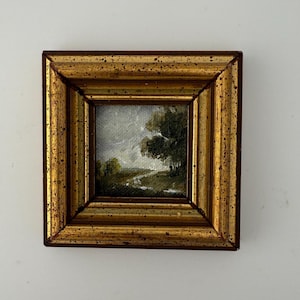 May include: A small square oil painting featuring a landscape with dark green trees and a winding path under a cloudy sky. The artwork is encased in a thick, ornate gold-toned frame with a speckled, distressed finish and dark brown edges.