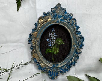 May include: An oval-shaped wall frame with an ornate, blue and gold painted finish. The frame contains a dark brown fabric backing featuring a small, embroidered botanical design of a light blue flower stalk with green leaves and stems.