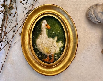 May include: An oval-shaped gold frame containing a small painting of a white duck with an orange beak and feet. The duck is depicted against a dark green background with small white floral details. The frame has a smooth, metallic finish.