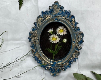 May include: An oval wall hanging featuring an ornate blue and gold frame with intricate scrollwork details. Inside the frame, a piece of black fabric serves as a background for a hand-stitched embroidery of three white daisies with yellow centers.