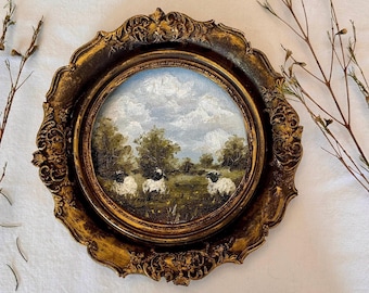 May include: A circular oil painting in an ornate, antique gold-toned frame featuring a landscape with three sheep grazing in a grassy field under a cloudy sky. The artwork is surrounded by thin, dried brown branches on a plain white fabric surface.