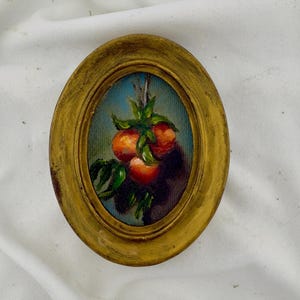 May include: An oval-shaped painting in a distressed gold-toned frame featuring a still life of three small orange fruits with green leaves hanging against a blue background. The artwork is rendered in oil paint with visible brushstrokes and texture.