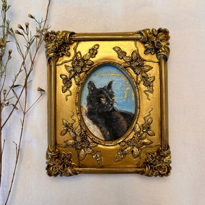 May include: A framed painting of a black cat with green eyes, set against a blue and gold background. The frame is ornate and gold with floral details.