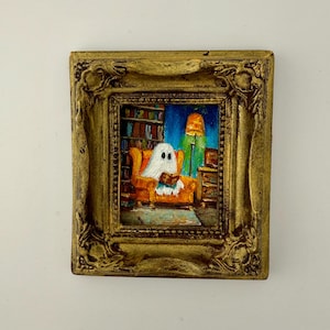 May include: A small framed painting shows a ghost reading in a living room. The ghost is in an orange armchair, with a bookshelf and a lit lamp behind. The frame is ornate and gold-coloured, adding a touch of elegance to the artwork. The painting is a whimsical piece.