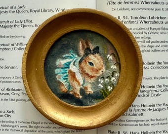 May include: A circular gold-toned frame containing a small painting of a brown rabbit wearing a blue ruffled garment. The rabbit is positioned next to white bell-shaped flowers against a dark background. The frame has a textured, matte finish.