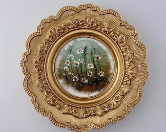 May include: A circular wall art piece featuring a painting of white wildflowers on a green and brown background, encased in an ornate, scalloped gold frame with intricate embossed detailing and a textured finish around the central circular border.