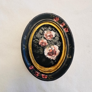 May include: A small oval painting of pink roses in a black and gold frame. The frame is decorated with small pink roses.