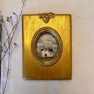 May include: A rectangular gold-toned frame with a textured finish and a decorative grape cluster at the top. The frame features a central oval opening containing a small oil painting of a white house with a brown roof under a cloudy sky.
