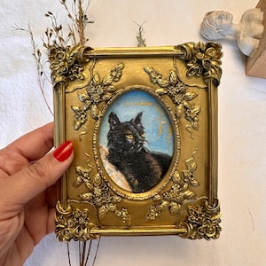 K&ouml;nnte beinhalten: Ein gerahmtes Miniaturgem&auml;lde einer schwarzen Katze mit gr&uuml;nen Augen vor einem blauen und goldenen Hintergrund. Der Rahmen ist verziert und golden mit floralen Details.