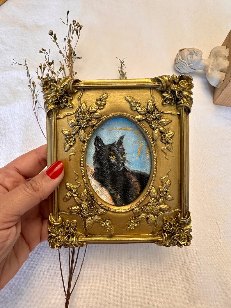 K&ouml;nnte beinhalten: Ein gerahmtes Miniaturgem&auml;lde einer schwarzen Katze mit gr&uuml;nen Augen vor einem blauen und goldenen Hintergrund. Der Rahmen ist verziert und golden mit floralen Details.