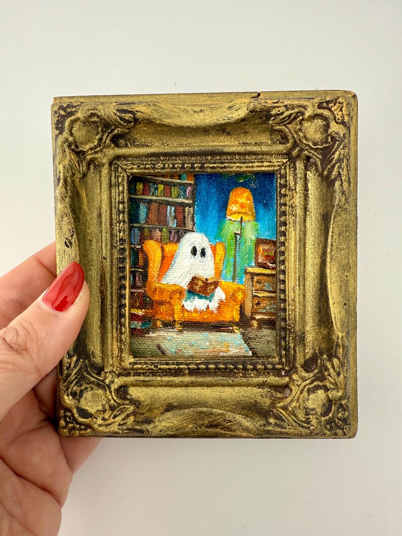 May include: A small, square painting in a gold ornate frame. The artwork depicts a ghost reading a book while seated in an orange armchair. A bookshelf, lamp, and side table are visible in the background. The painting has a whimsical, cozy feel.
