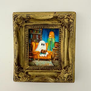 May include: A framed miniature painting depicts a ghost reading in an orange armchair in a library. The painting has a gold ornate frame. The background shows bookshelves, a lamp, and a blue sky.