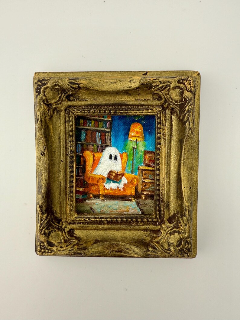 May include: A framed miniature painting depicts a ghost reading in an orange armchair in a library. The painting has a gold ornate frame. The background shows bookshelves, a lamp, and a blue sky.