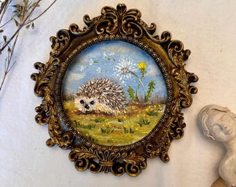 Puede incluir: Una pintura al óleo circular de un erizo en un campo con dientes de león, encerrada en un marco dorado ornamentado de estilo antiguo con intrincados motivos de volutas y hojas. La pintura muestra un cielo azul, hierba verde y flores amarillas.