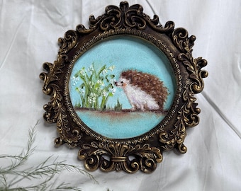 Puede incluir: Una pieza de bordado circular que presenta un erizo marrón y blanco junto a flores blancas sobre un fondo azul claro, encerrada en un marco ornamentado de color bronce oscuro con volutas intrincadas y adornos decorativos.