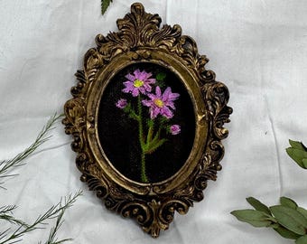 May include: An oval-shaped, ornate gold-colored frame featuring intricate scrollwork and relief details. Inside the frame is a black fabric background displaying a hand-stitched embroidery of two purple flowers with yellow centers and green stems.
