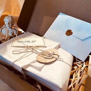 K&ouml;nnte beinhalten: Eine wei&szlig;e Schachtel, die in braunes Papier eingewickelt ist, mit einer Schleife aus Schnur und einem kleinen Etikett mit der Aufschrift "Miss Lily Art", liegt in einem braunen Karton mit Stroh als F&uuml;llmaterial. Ein wei&szlig;er Umschlag mit einem Wachssiegel befindet sich ebenfalls in der Schachtel.