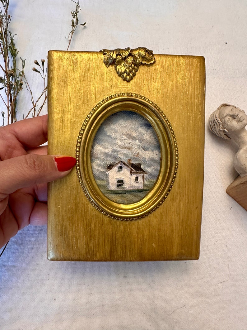 May include: A rectangular gold-painted wooden frame featuring an oval center cutout that displays a small oil painting of a white house under a cloudy blue sky. The frame is adorned with a decorative metallic grape leaf accent at the top center.