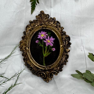 May include: A framed embroidery of a single pink flower with green leaves on a black background. The frame is gold and ornate.