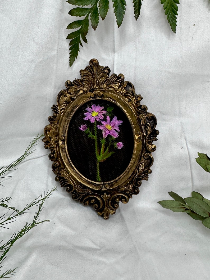 May include: A framed embroidery of a single pink flower with green leaves on a black background. The frame is gold and ornate.